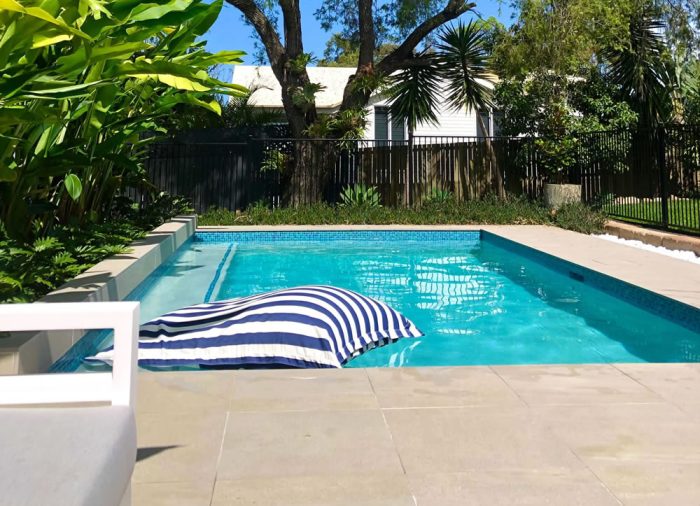 Beautiful aqua blue backyard swimming pool with a blue and white striped pool lounge, surrounded by lush green trees