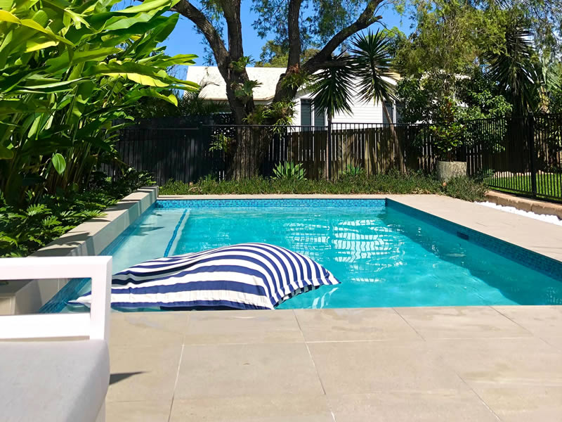 Beautiful aqua blue backyard swimming pool with a blue and white striped pool lounge, surrounded by lush green trees